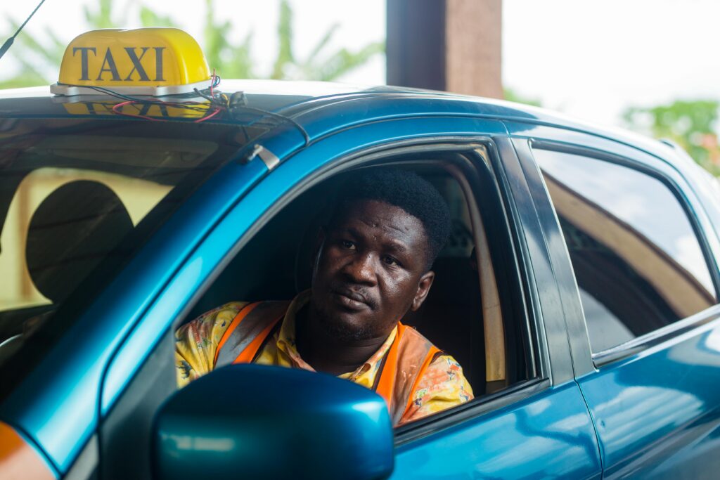 A taxi driver sitting in a blue car, wearing an orange vest, waiting for passengers.